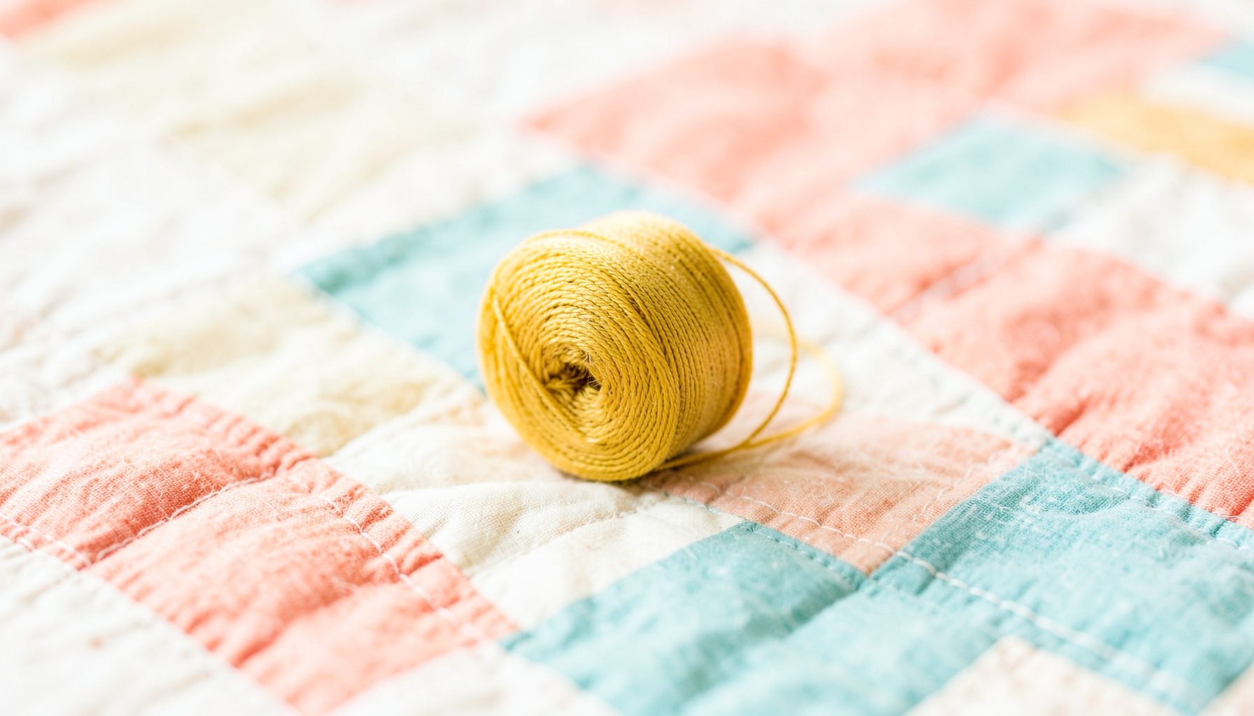 Spool of golden-yellow thread resting on a patchwork quilt with soft cream, coral, and teal fabric squares, warm and inviting craft atmosphere