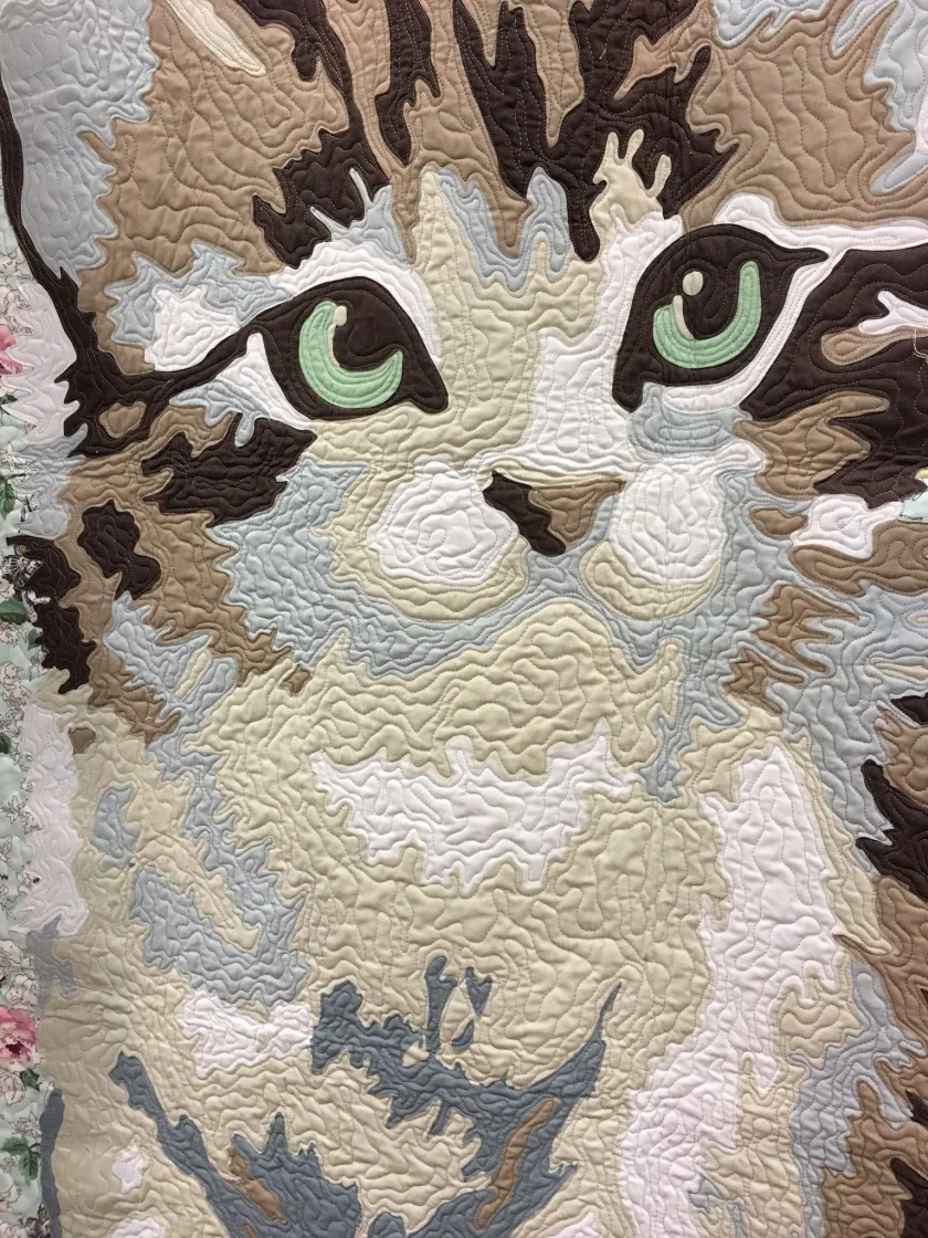Close-up of a quilted textile artwork depicting a cat's face in beige, brown, and cream tones with green eyes, soft handmade feel