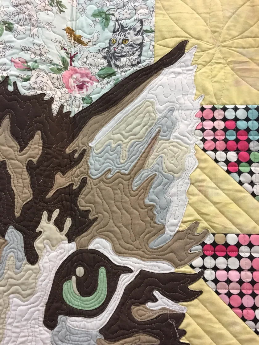 Close-up of quilted fabric showing an appliqued bird wing in brown and cream tones next to patterned fabric squares, warm soft light