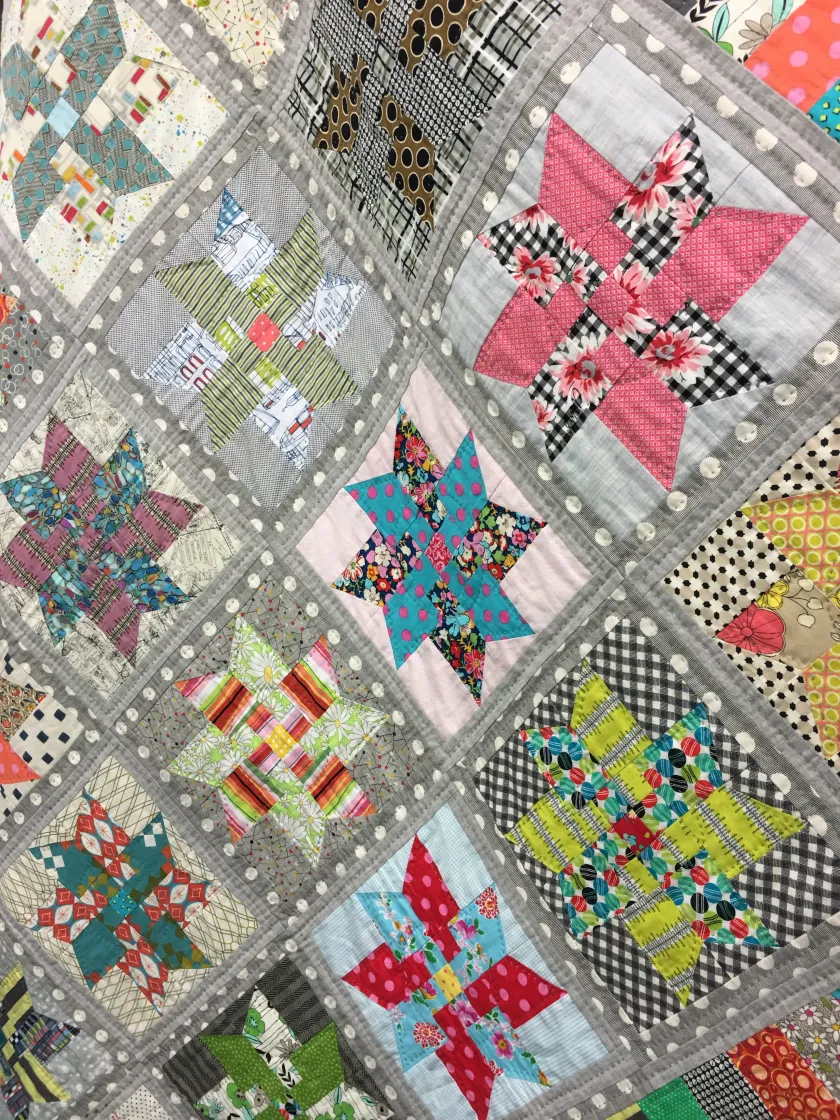 Patchwork quilt featuring star-shaped blocks in a variety of bright floral and graphic fabrics bordered by gray polka-dot sashing