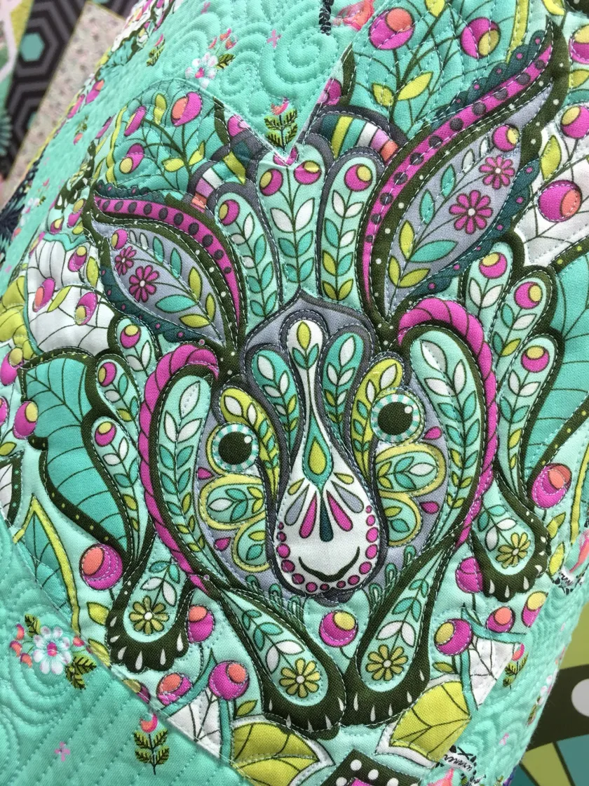 Close-up of a quilted textile with an ornate floral and paisley-style animal face design in teal, pink, green, and gray tones