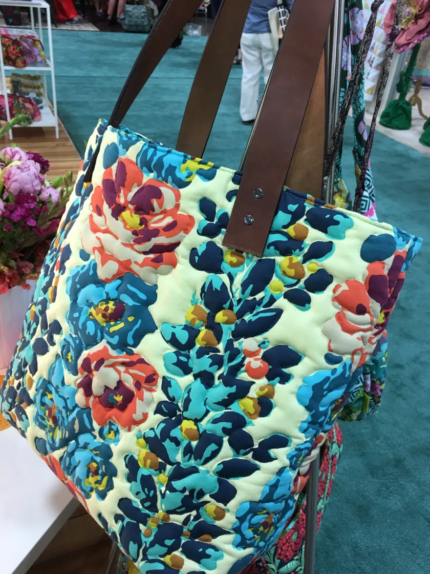 Quilted tote bag with floral pattern in teal, orange and cream tones with brown leather straps, hanging indoors near flooring