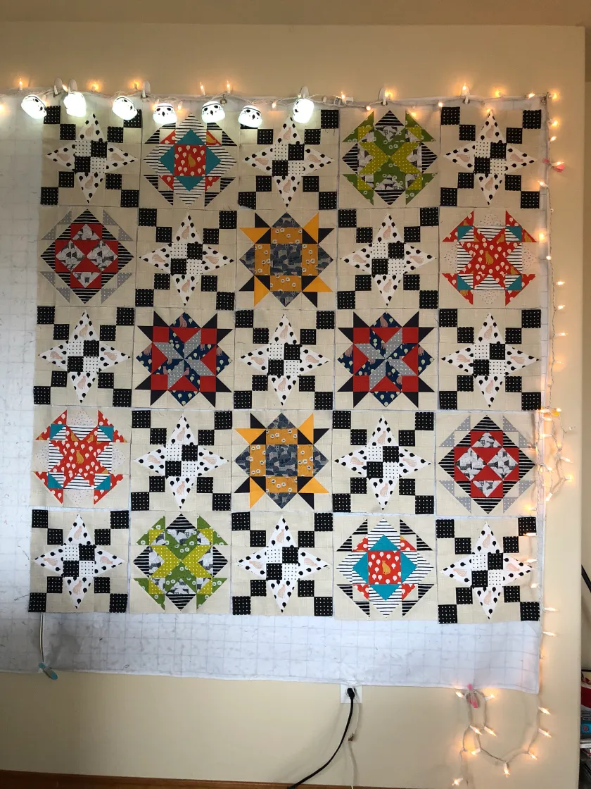 Patchwork quilt top with star and checkerboard pattern in red, yellow, green, and black and white, hung on wall framed by warm wood tones